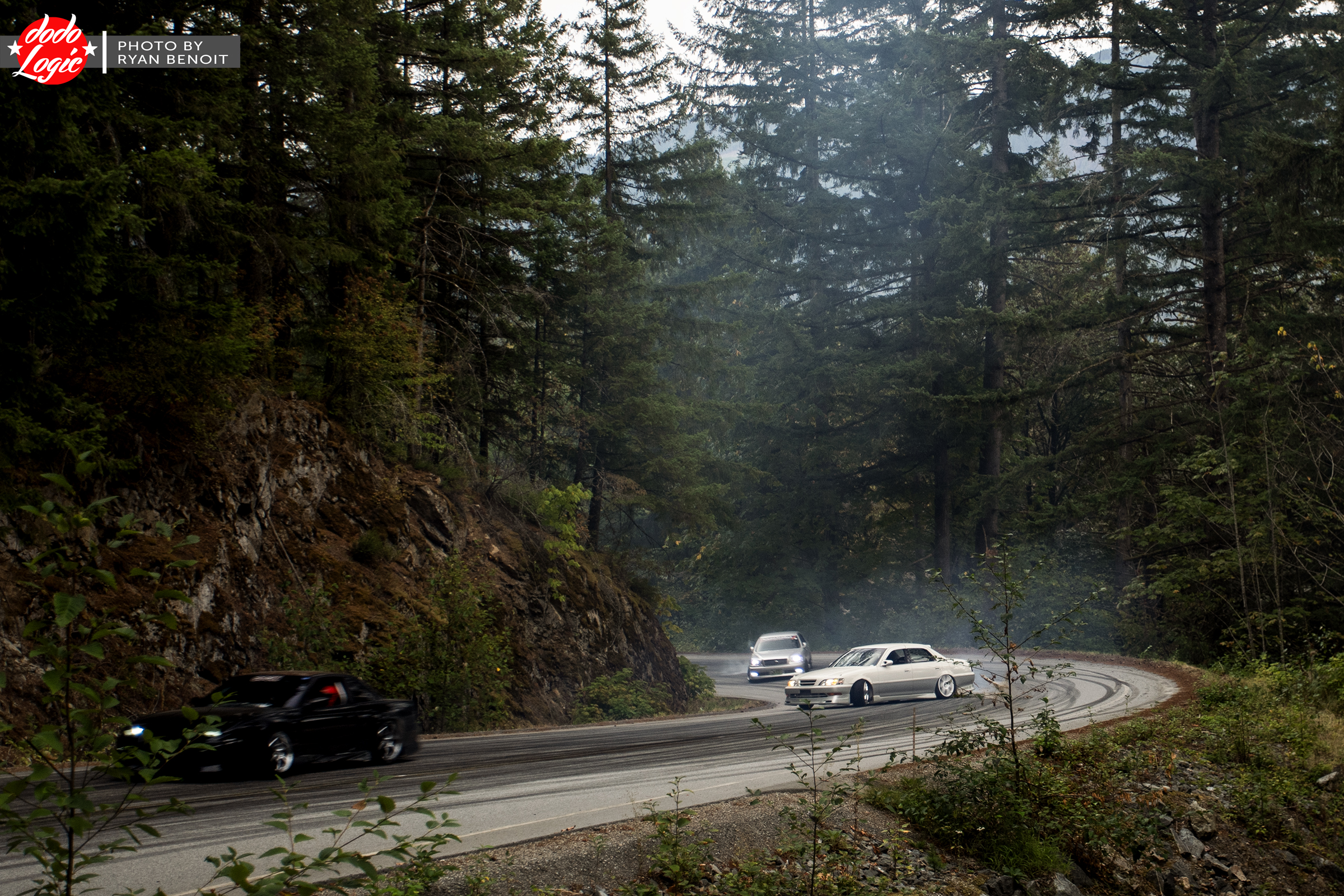 Two distant cars wind around a forest road as tire smoke hangs among the trees
