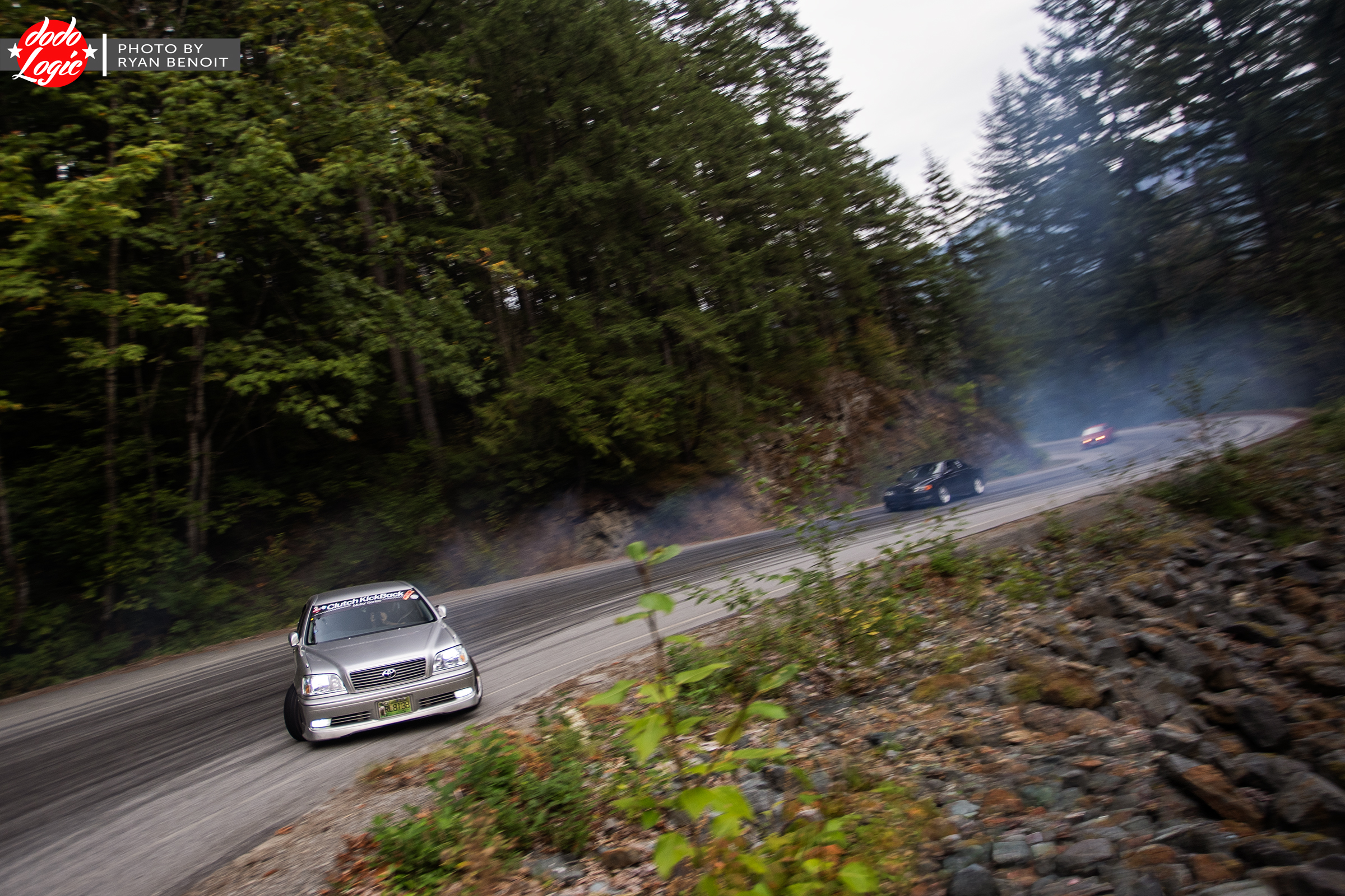 Cars appear around a distant mountain bend beyond a rocky shoulder