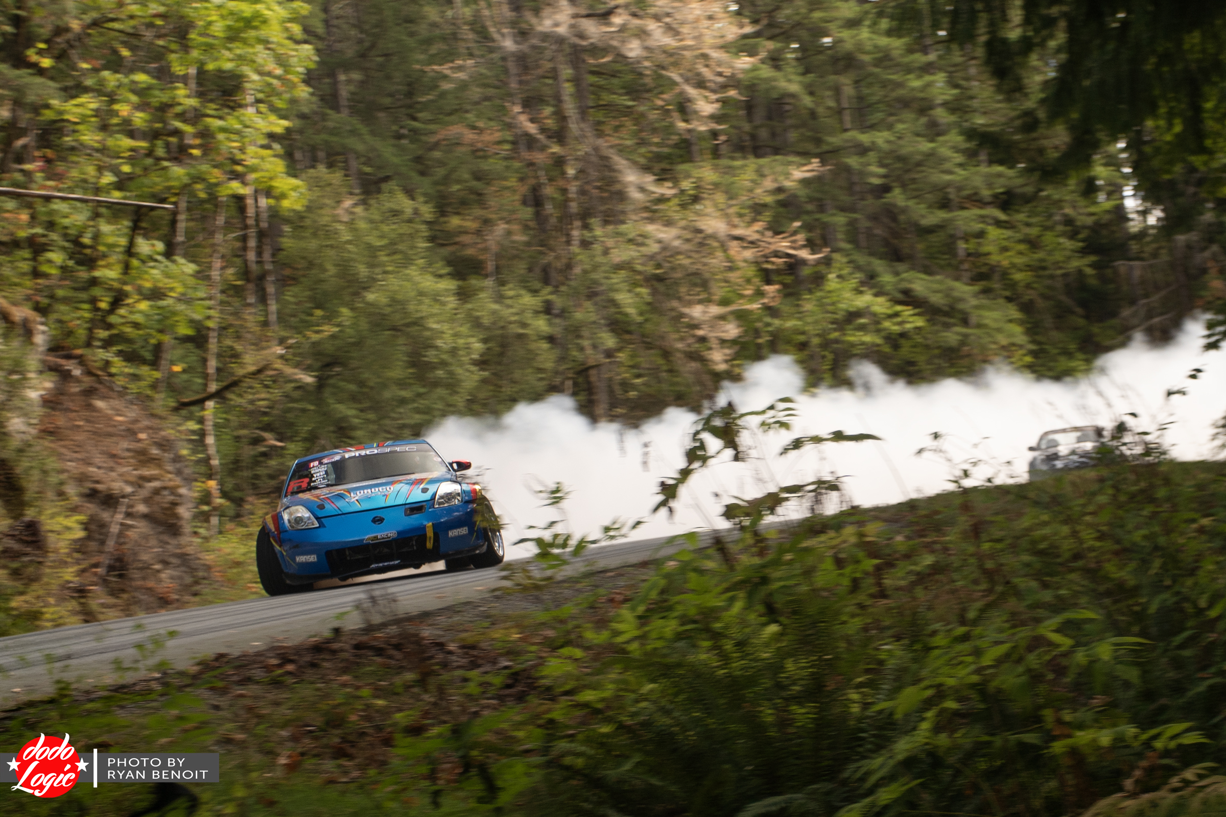 Blue drift car crosses a wooded mountain road trailing a long plume of tire smoke